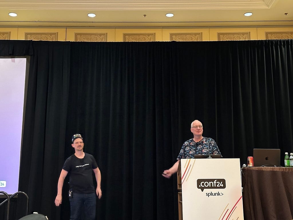 Two individuals are on stage at a .conf24 Splunk conference, with one standing behind a podium and the other nearby.