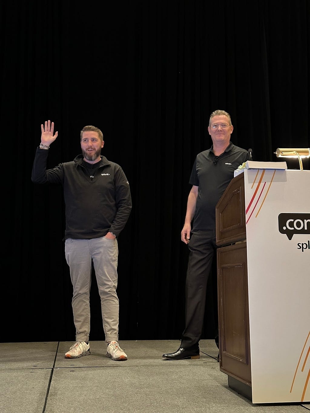 Two men stand on a stage with one waving, beside a podium with a Splunk logo.
