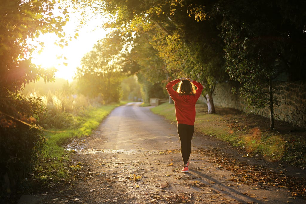 A woman walks on a paved pathway with her hands on top of her head. She is wearing an orange or red sweatshirt with black pants, and she is positioned in the foreground walking away from the camera. The sun is shining into the picture from the upper left corner and the path she walks is lined with trees as well as green grass. There are a few dead leaves on the trail around her.