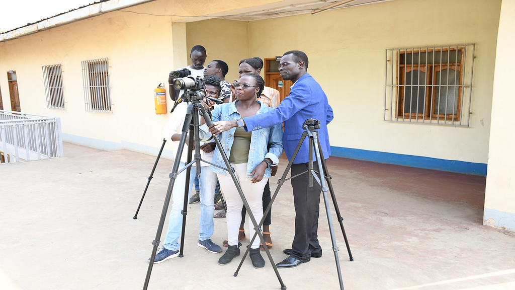 A group of six journalists stand around a camera on a tripod as part of a practice section to improve their videography skills.