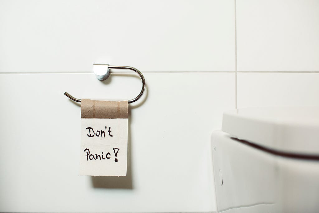 One last, lonely square of toilet paper hangs from the barren cardboard roll in a bathroom, with the words written upon it: “Don’t Panic!”