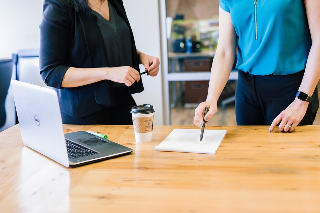 Two people standing at a desk with laptop, coffee, pen and paper on desk / Photographer: Amy Hirschi | Source: Unsplash