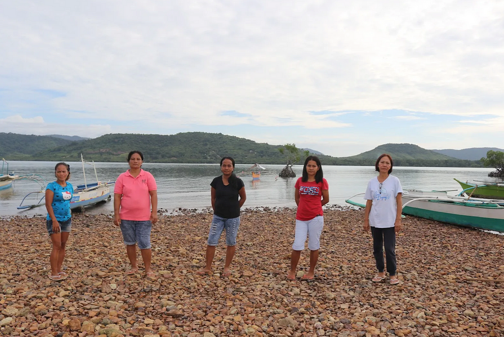 Five women stand on a shoreline in front of a lake with several boats.