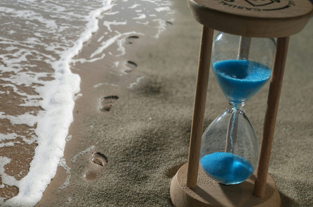 An hourglass sitting in the sand on a beach with footprints by the waters edge