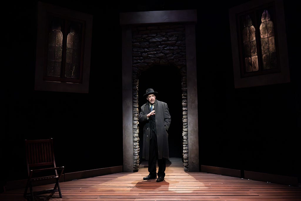 A man in an overcoat and hat stands in a stone archway between two old windows