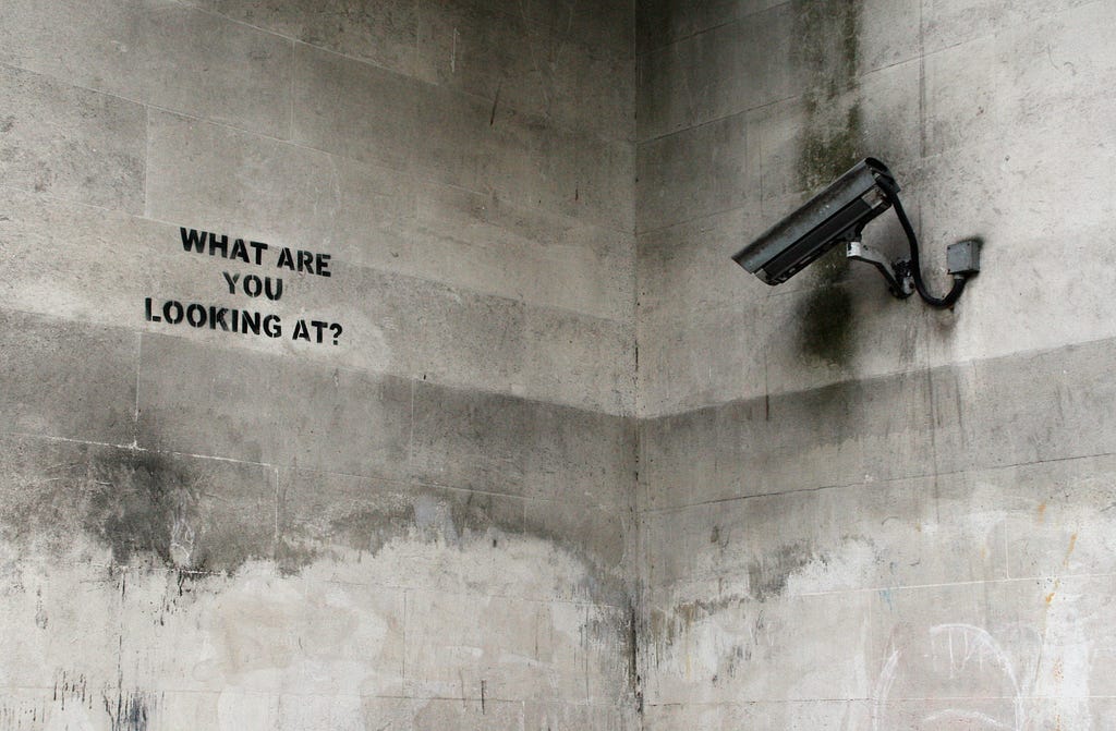 a photo of a security camera pointing at the phrase “what are you looking at” painted on a wall