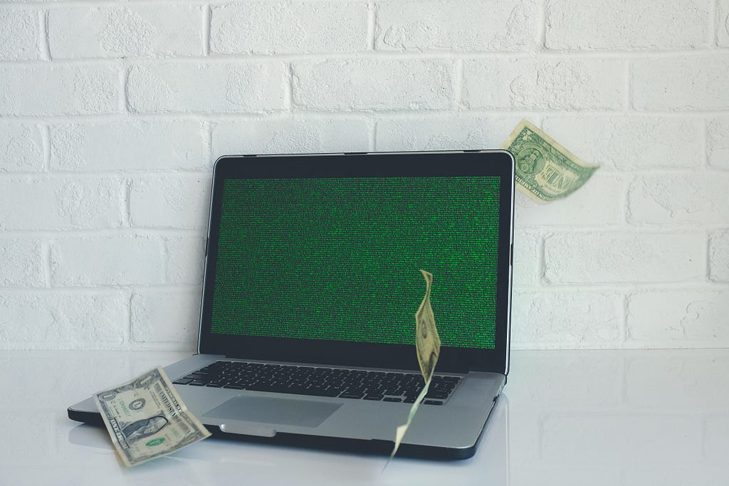 A laptop computer sits on a counter with three 1 dollar bills falling onto it from above.