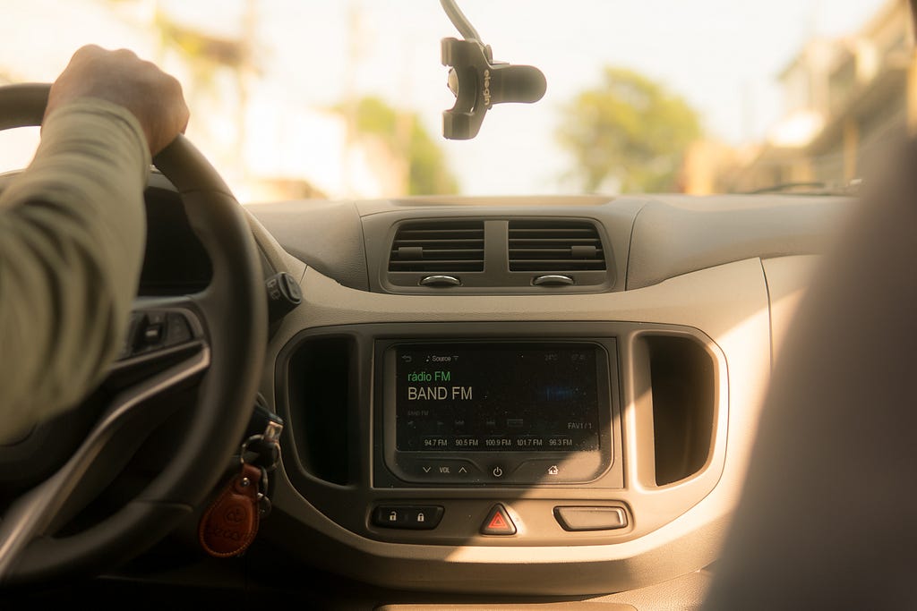 A digital display on the front dashboard of a car indicates “BAND FM” radio is playing. Golden sunlight shines through the windshield. A man’s arm grips the steering wheel. He is wearing a tan-green sweatshirt. Blurry trees can be made out through the windshield in the background.