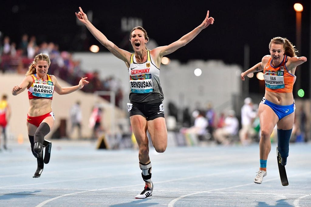 3 women with lower leg amputations and wearing blades running in a race, the middle woman is ahead and cheering with arms spread wide