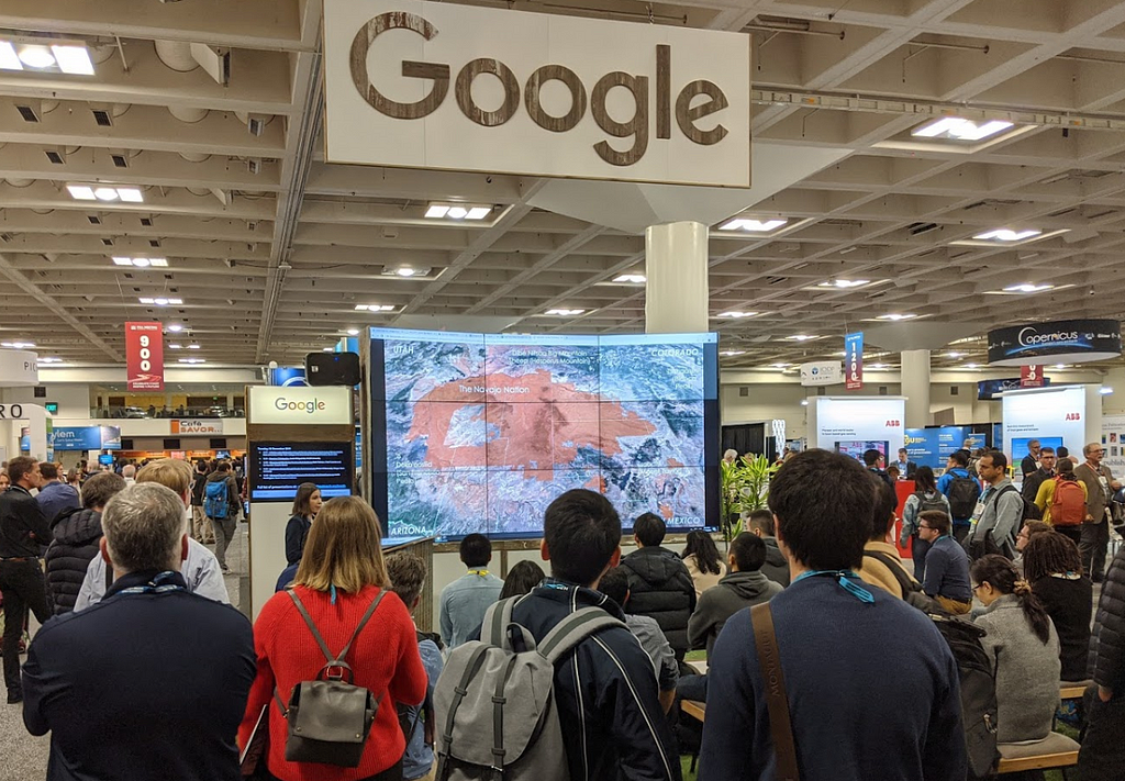 Photo of 2019 Google booth lightning talks in exhibit hall, with a large monitor displaying a Google Earth map from a speaker.