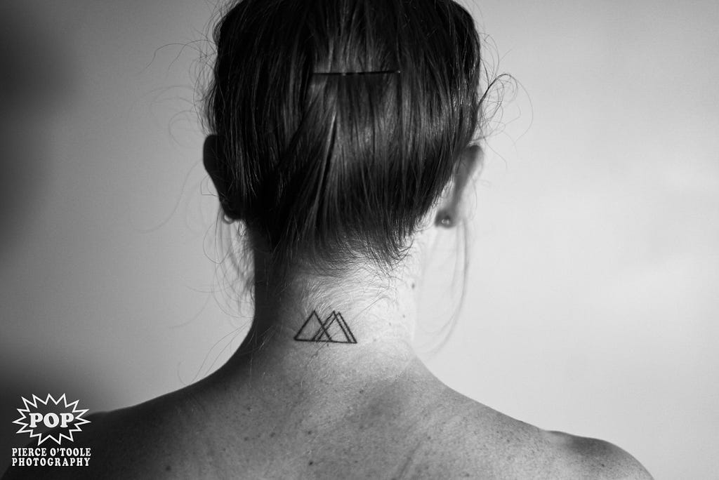 black and white photo of the back of woman from shoulders to head with a triangle tattoo on her neck