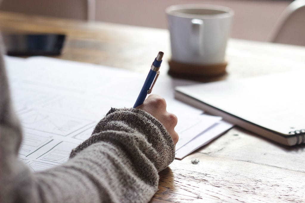 Photo of a person sketching in a notebook next to a cup of coffee.