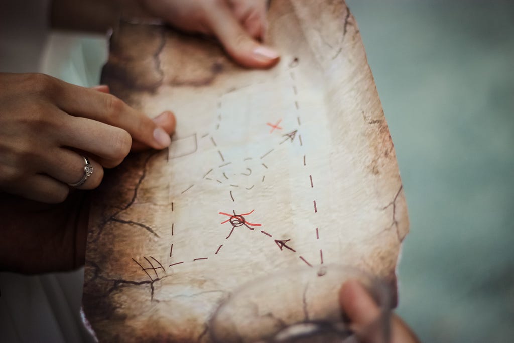 Two sets of hands holding a hand drawn treasure map. One of the hands is pointing to a position on the map.