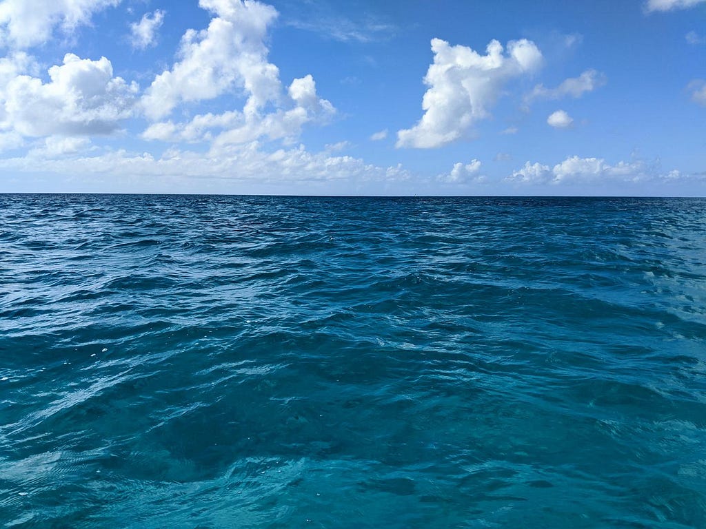 The open ocean, with blue sky above and white clouds on the horizon