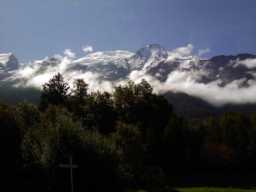 photo of snow-covered alps jutting up above some low clouds, with green trees and a tiny road sign in the foreground