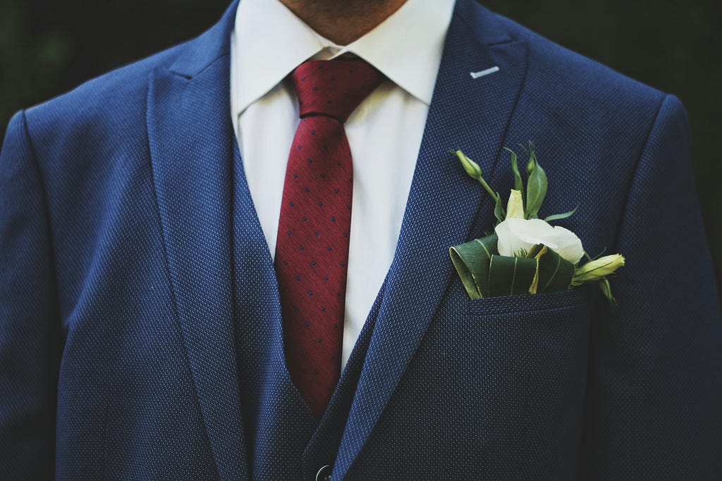 A groom in a blue tuxedo with flower in the breast pocket.