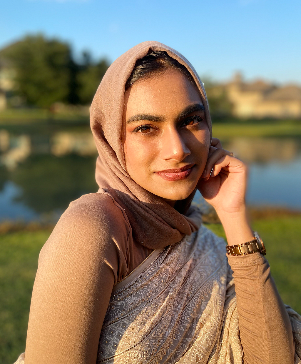 Anika poses in front of a lake on a sunny day, wearing a beige hijab and matching top.