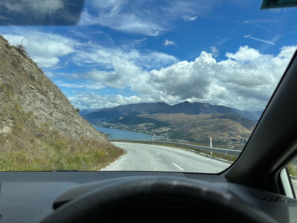 View from driver’s seat of a right-hand drive car with top of steering wheel visible. Lake, mountains and sky visible through the windshield.