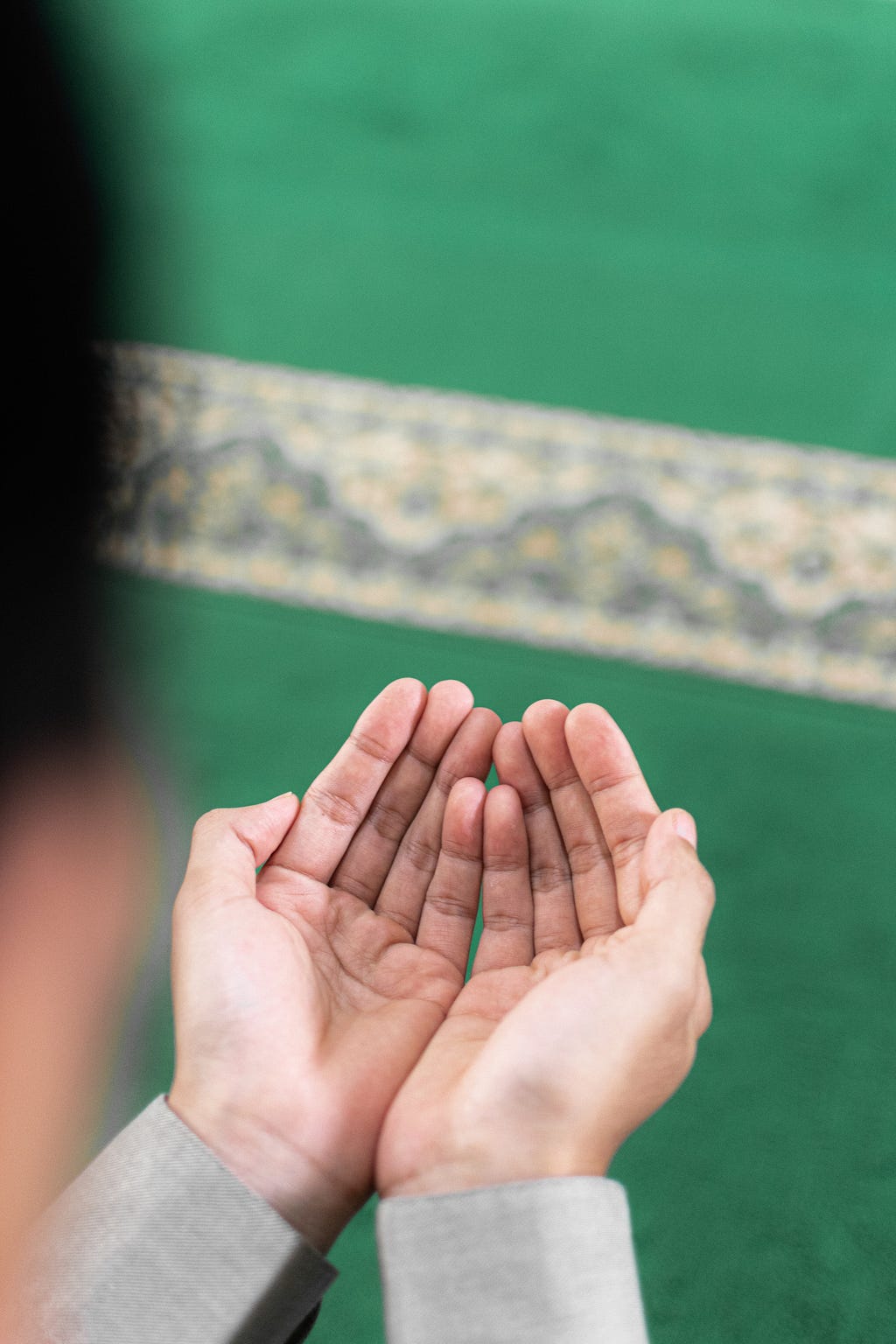 Two hands cupped in prayer on a green background of a rug with an indistinct woven border slashing diagonally along the top of the picture.