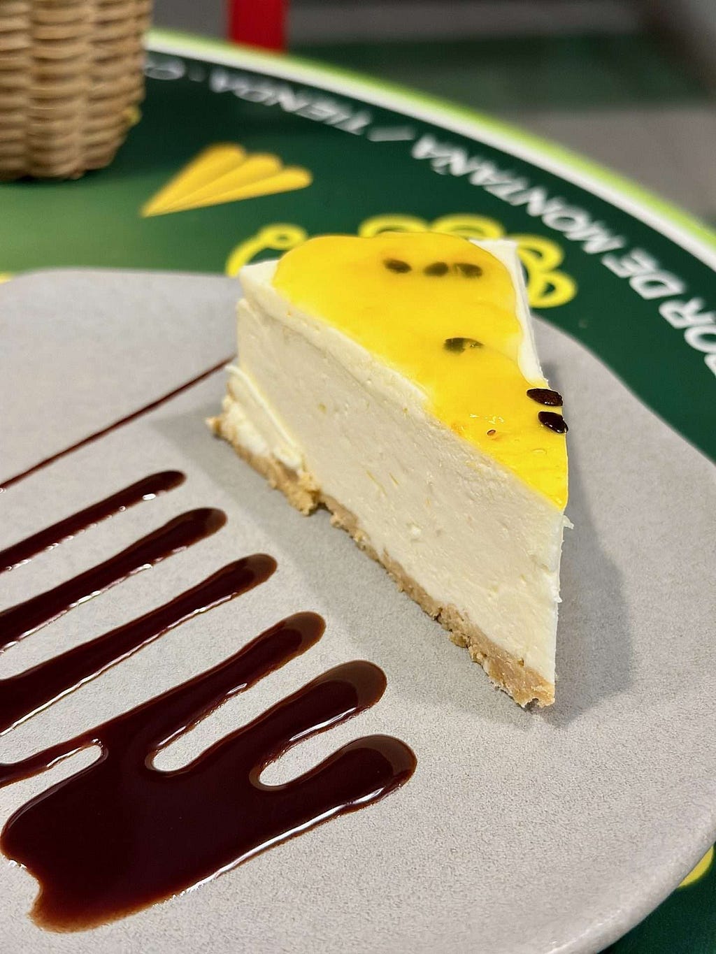 A slice of cheesecake with yellow passionfruit topping, served on a gray plate with chocolate sauce drizzled beside it. A delicious Cartagena, Colombia dessert