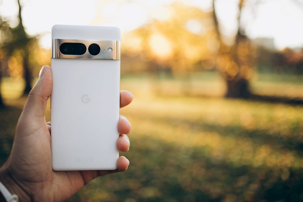 A Google Pixel 7 Pro being held in focus against a blurry background of a field with bright sun in the sky.