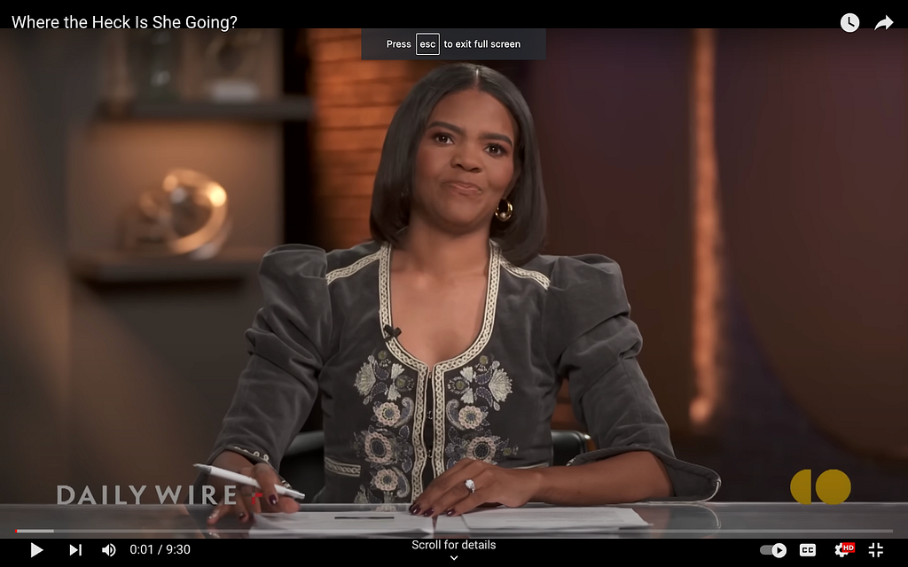 A screenshot from Candace Owens’s show on The Daily Wire. What she’s wearing might be a dress or a type of blazer, since we only see her from the waist up as we did in all the previous screenshots. It has a scoop neckline with a silvery trim. It’s black and looks like it’s made of suede. The shoulders have a slight puff and the front features embroidered botanicals in the same silver as on the neckline.