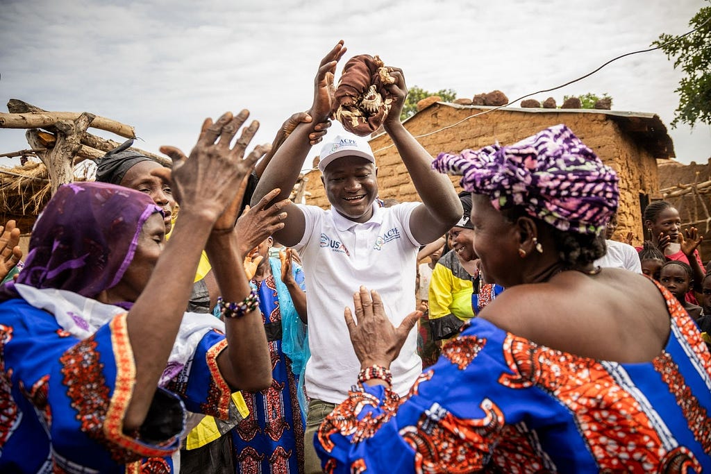A smiling man holding his arm aloft is surrounded by women in colorful dresses who are clapping and smiling.