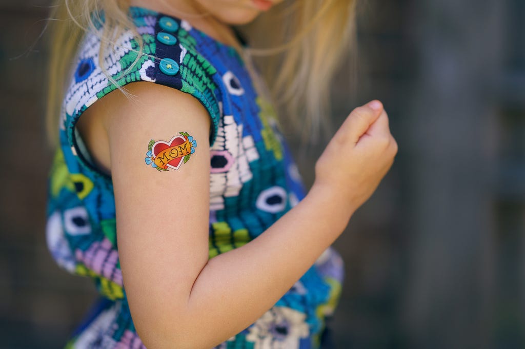 Partial side view of a young girl in a floral dress with a with a temporary “mom” tattoo just under the shoulder.