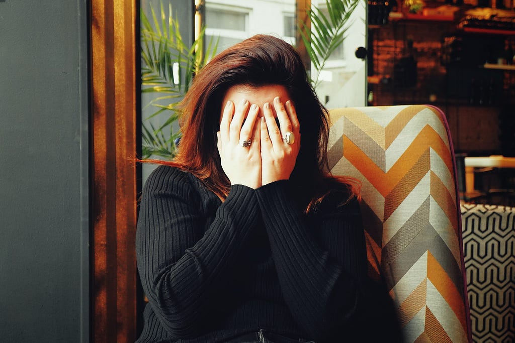 Woman with dark hair and black sweater sits in chair in front of window, covering her face with both hands.