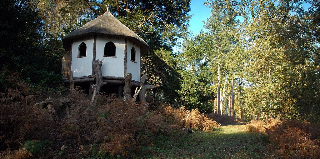 A small hut with white walls and a thatched roof standing on log stilts in a forested area.