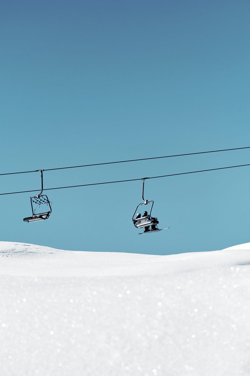 Ski lift chairs against a clear blue sky, with two skiers on the further chair above a sparkling snow-covered slope. The descending chair is empty.