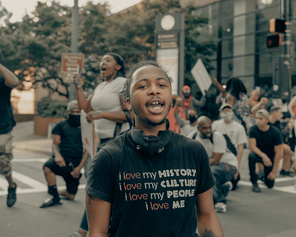 Photo of a young African American boy at a protest wearing a t-shirt that says: “I love my history, I love my culture, I love my people, I love me.”