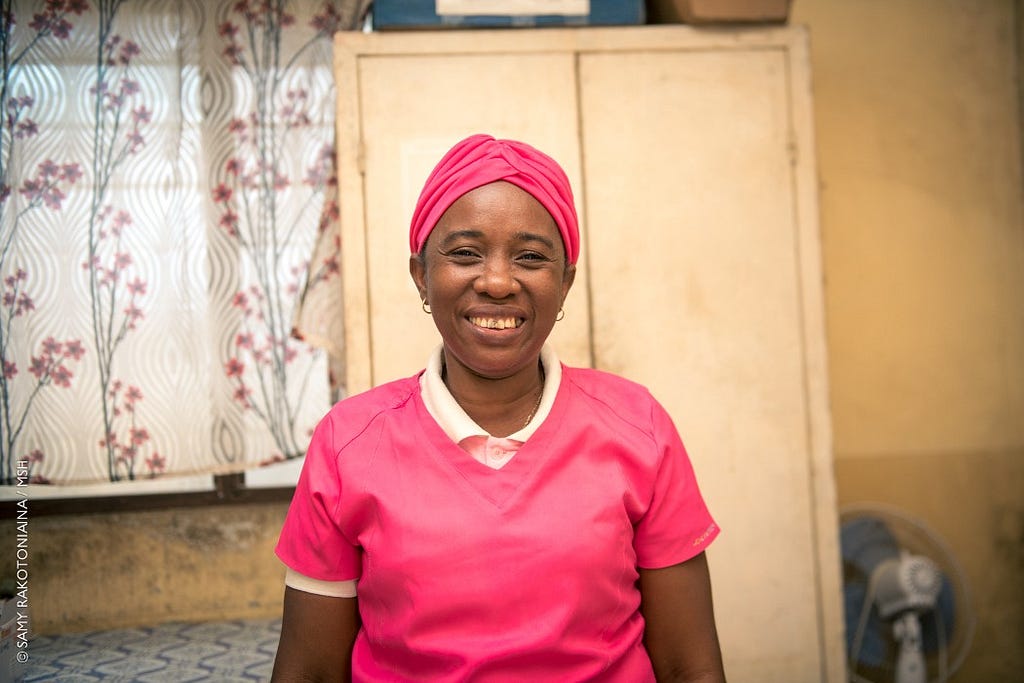 A Malagasy midwife in pink scrubs and a pink headscarf smiles for the camera.