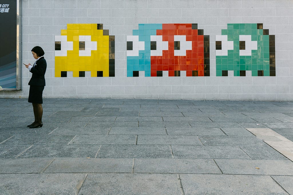 Asian woman uses a mobile device. On the wall in the background, four Pac-Man ghosts (yellow, blue, red, green) appear to be sneak peeking from behind her.