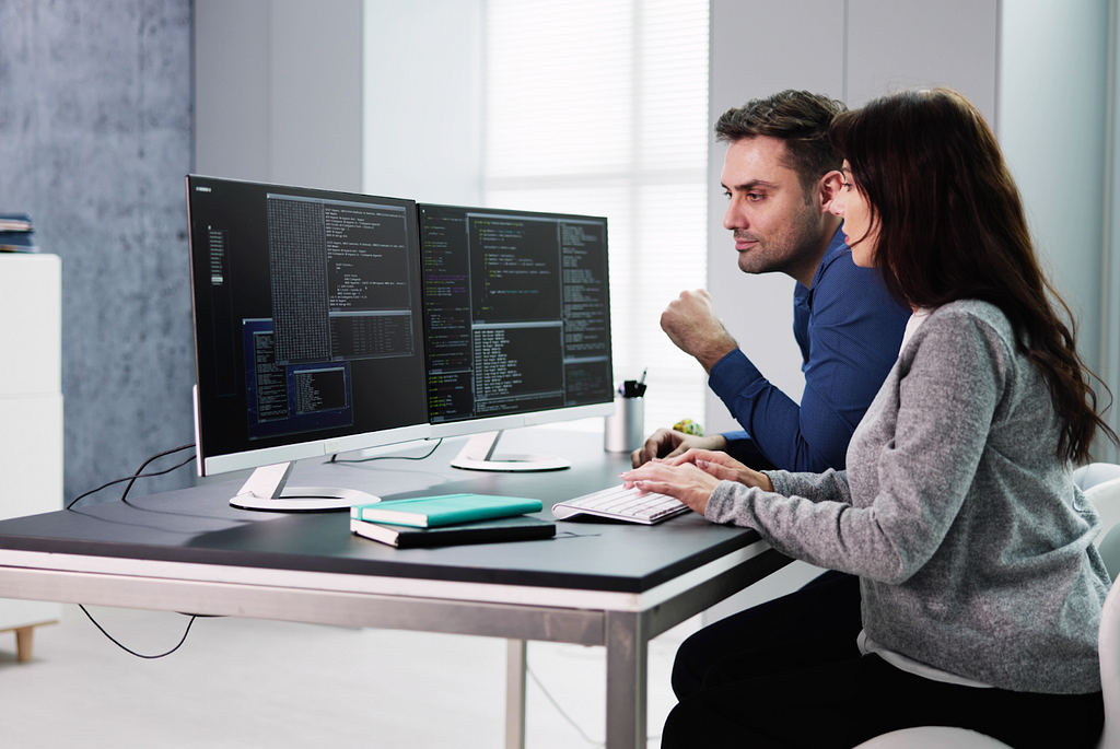 Two coworkers, a woman and a man, collaborate while running code on two computers. They engage in a focused discussion about Feature-Driven Development (FDD), emphasizing teamwork and analytical problem-solving in a modern office setting.