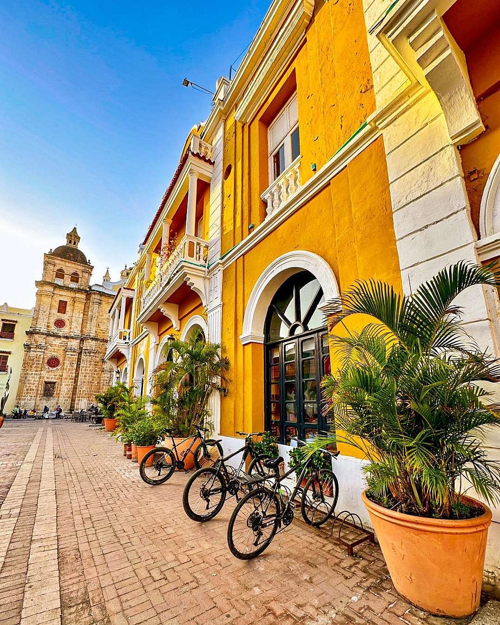 A row of yellow and white buildings with arched windows, potted plants, and bicycles lined up on a cobblestone street in Cartagena, Colombia, with a stone tower in the background, under a clear blue sky.
