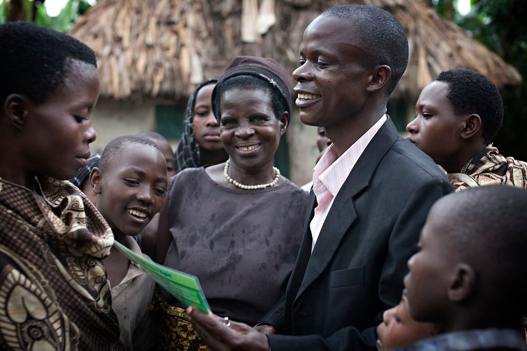 A group of health cooperative members in Uganda smile, looking towards a piece of paper held by a man in the middle.
