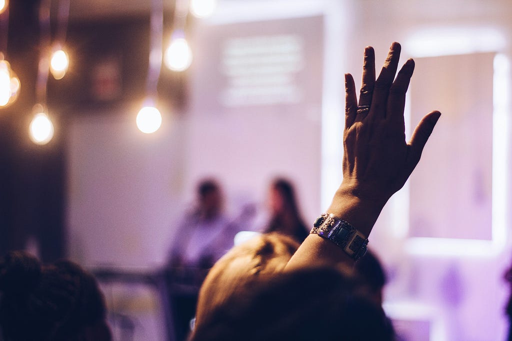 Blurred image from what seems to be a meeting or class room, where one person raises the hand to ask a question