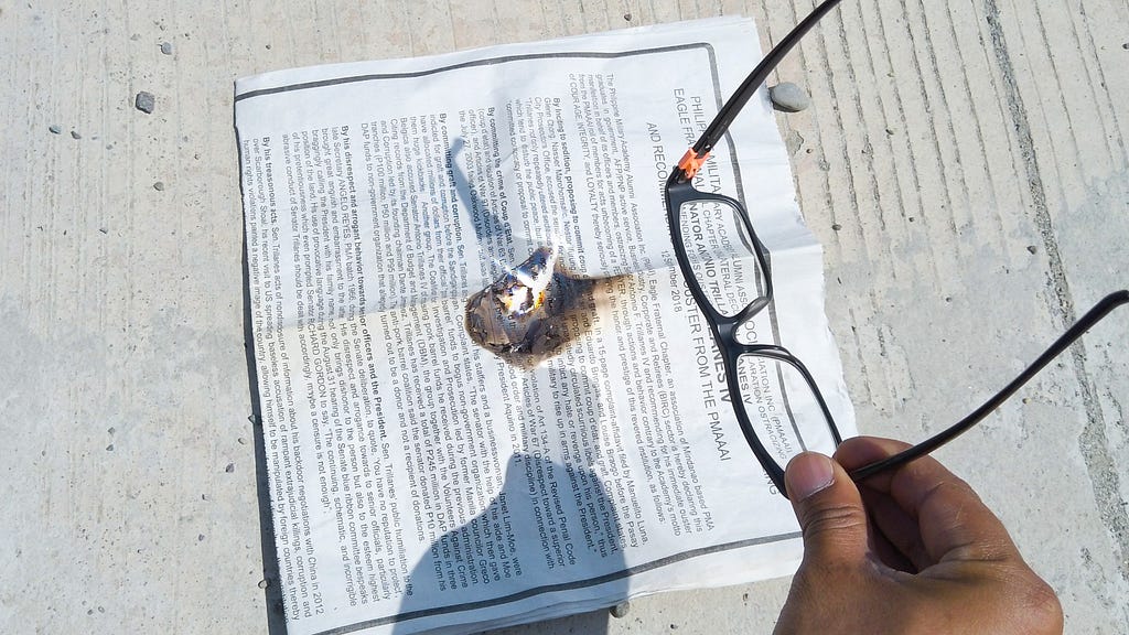 A pair of glasses are held under direct sunlight to start a fire on a newspaper.