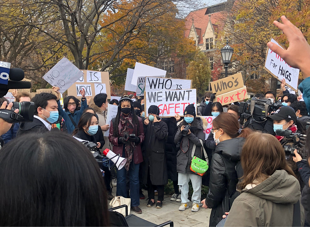 A group of people of various ages and gender presentations, mostly East Asian, gather in a loose circle around a young woman holding a megaphone. Signs held by the crowd read “Who is Next?” and “We Want Safety.”