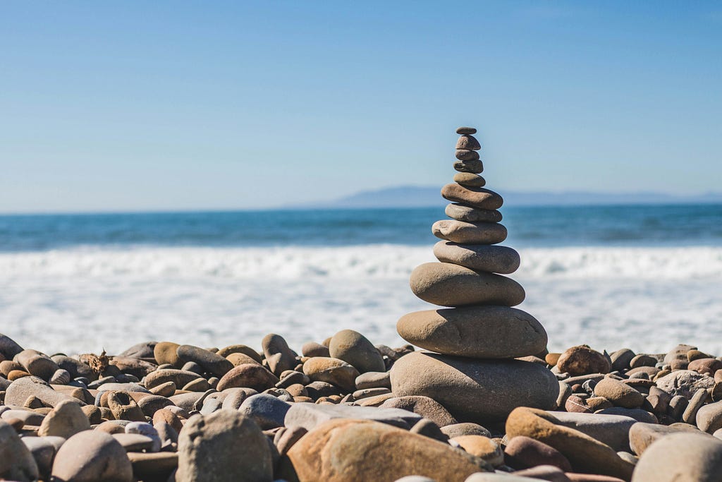 Na imagem, rochas e pequenas pedras perfeitamente equilibradas estão em primeiro plano. Ao fundo, as ondas e o mar em um vasto horizonte praiano.