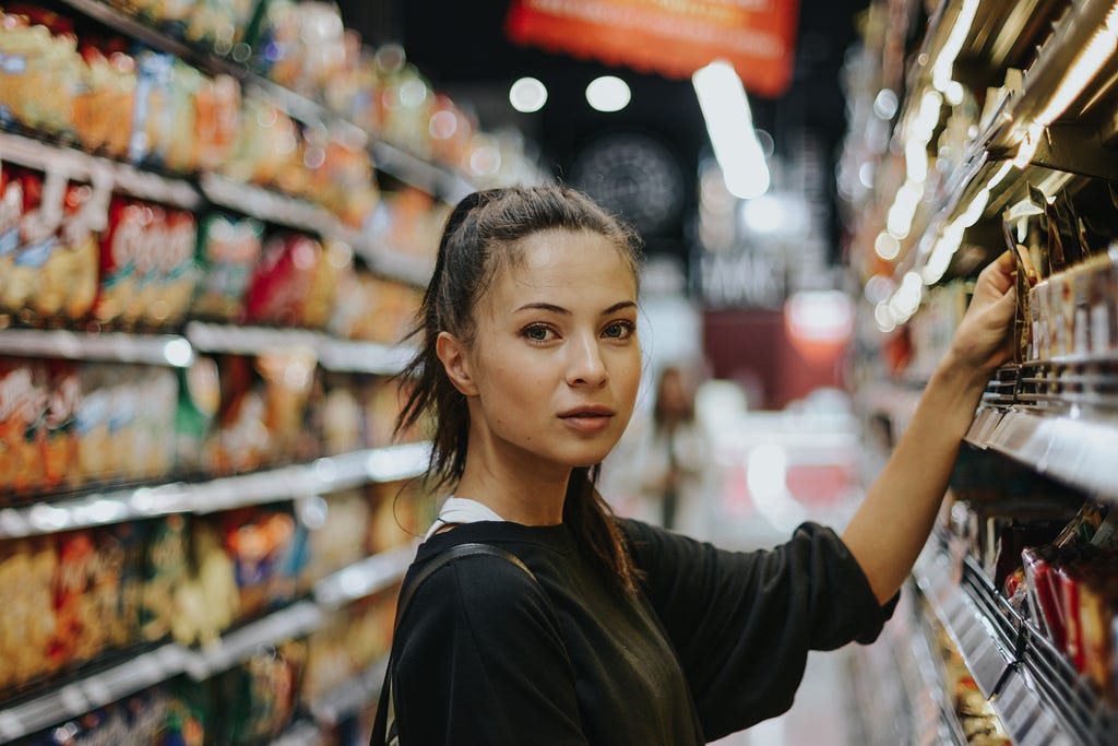 A woman with light skin and dark hair in a pontytail lifting her arm to pick up an item from the shelf at the grocery store.