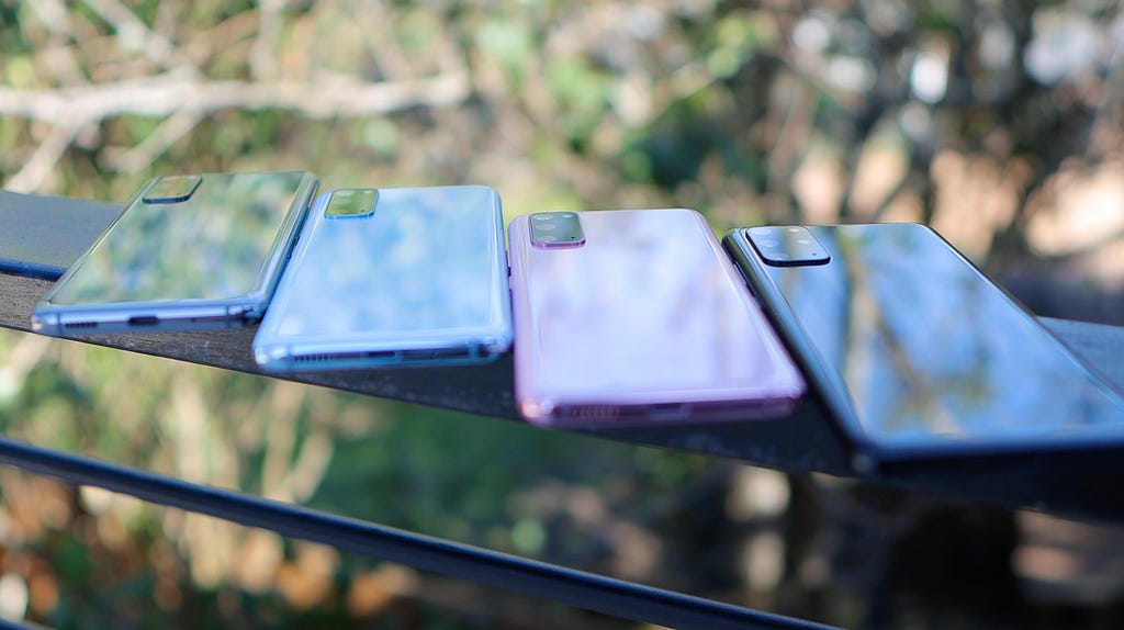 Four smartphones in different colors sit face-down on a glass table. On their cases and the table, you can see the reflections of trees and the sky.