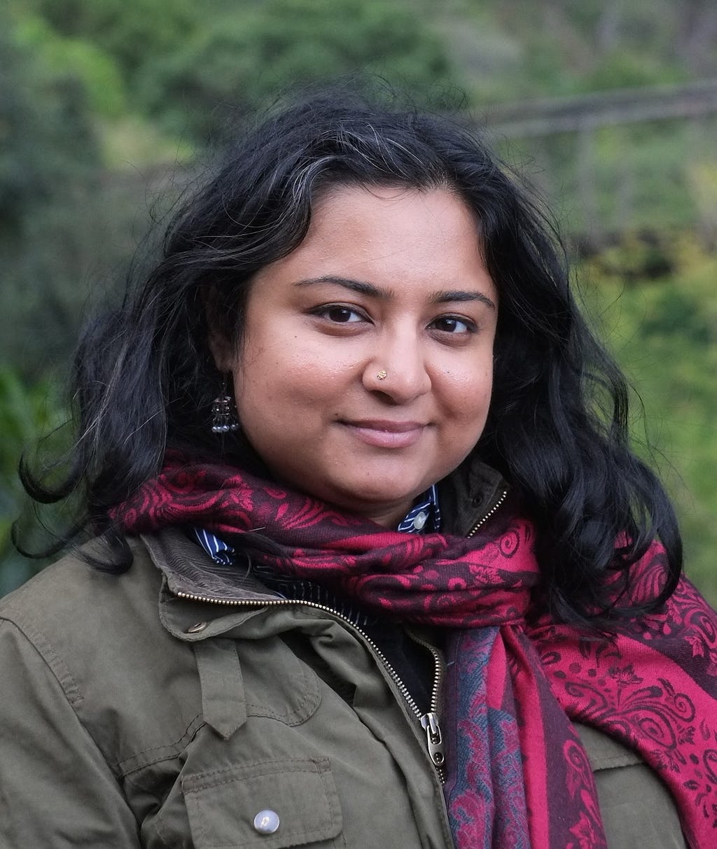 Choudhry in an olive jacket, dark magenta patterned scarf, nose ring and earrings, greenery behind.