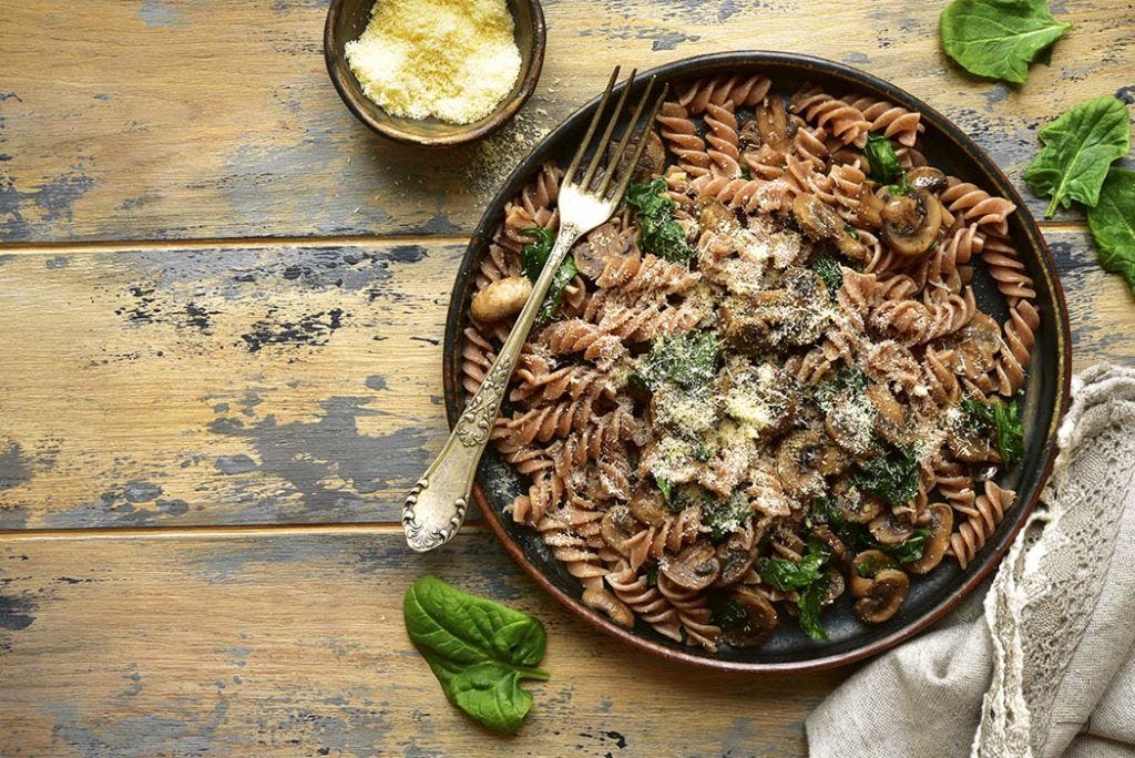 Spinach and mushroom whole wheat pasta topped with freshly grated parmeson cheese in a brown bowl, on a wooden table.