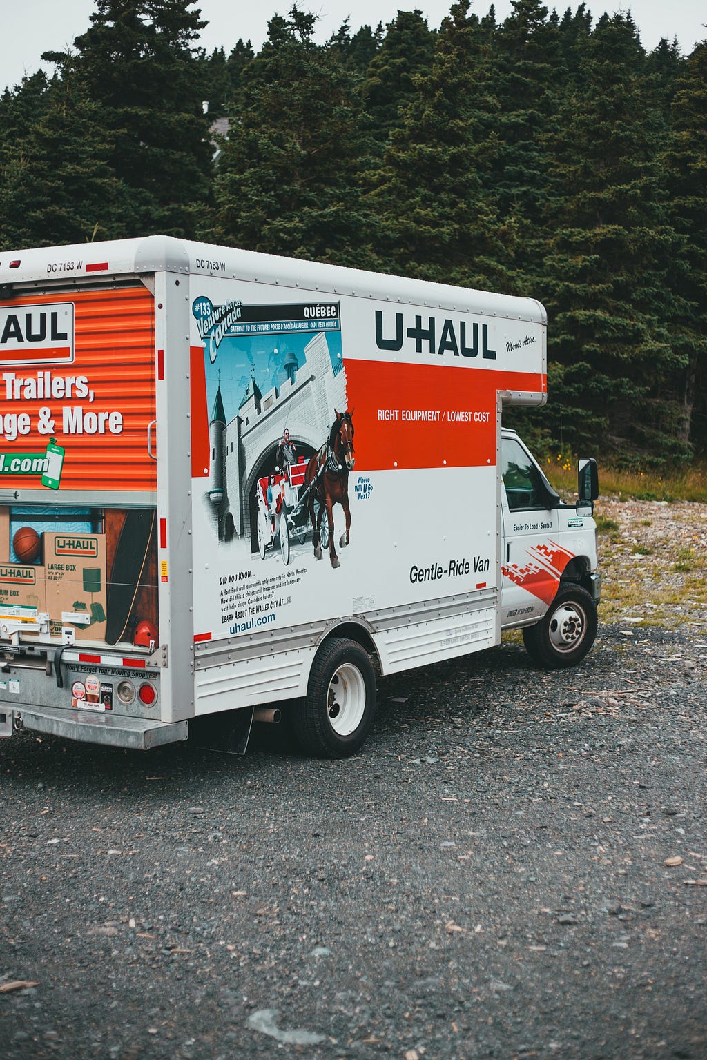 A unhaul truck pulled to the side of a road on gravel. Behind it are pine trees and an afternoon sky