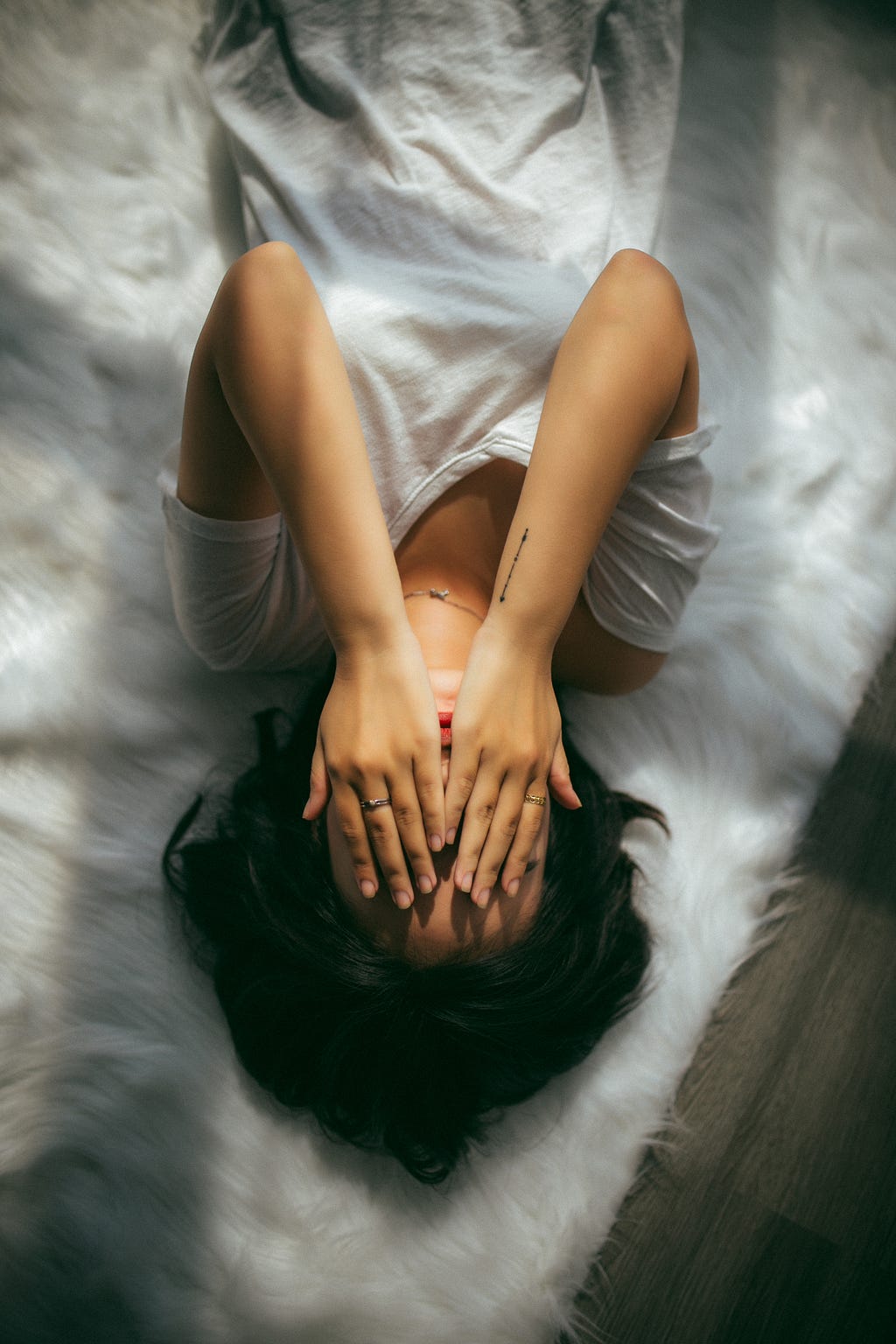 A picture of a women wearing a white t-shirt, laying on her bed, while covering her face with her eyes. Her bed was covered with some kind of an animal’s fur rug. The picture was taken upside-down, so the head is on the bottom of the picture.