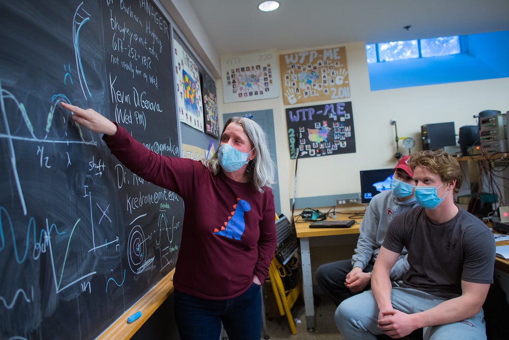 A woman wearing a mask stands at a blackboard explaining a mathemetical concept to two masked, male students