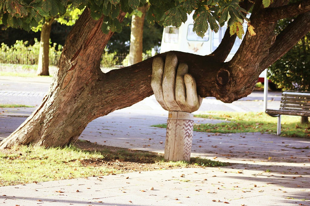 A large tree branch leans heavily to the side and is supported by a sculpted stone hand emerging from a column. The hand gently cradles the branch, symbolizing external support holding hidden weight. The scene takes place in a quiet, sunlit park.