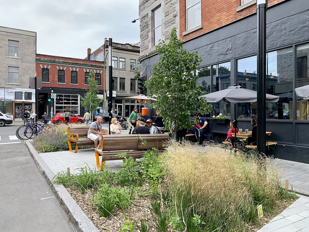 The same street corner, previously with no greenery, now with an extended sidewalk with seating, trees, and other landscaping.